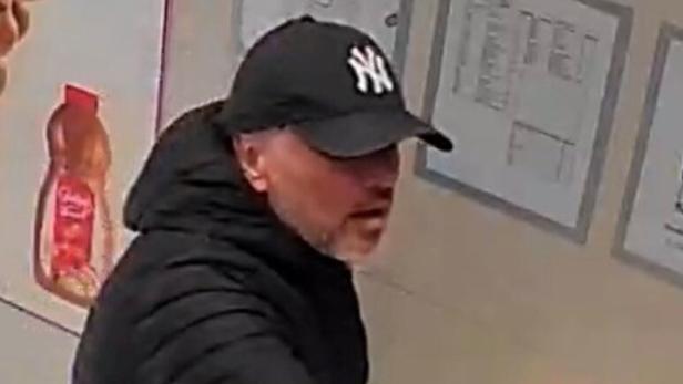 Mann in schwarzer Jacke und New-York-Cap zeigt mit ausgestrecktem Arm auf etwas außerhalb des Ausschnitts.