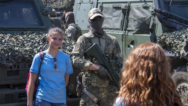 Ein bewaffneter Soldat in Tarnuniform steht vor Militärfahrzeugen, daneben ein lächelndes Mädchen in blauem Shirt.