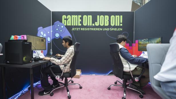 Zwei junge Männer spielen an PCs auf einer Gaming-Messe mit großem „Game on. Job on!“-Banner.