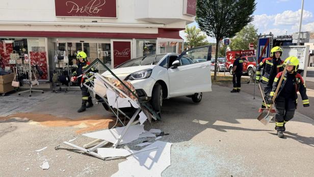 Auto prallt gegen zerstörtes Ladenschild vor einem Geschäft; Feuerwehrleute sichern die Unfallstelle.