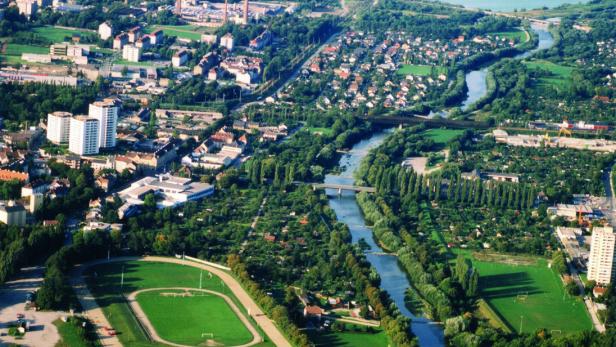 Luftaufnahme einer Stadt mit Fluss, vielen Grünflächen, Wohnhäusern und Feldern am Rand.