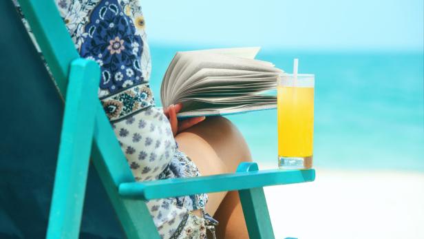 Woman sitting in deck chair at beach reading book on sunny day.