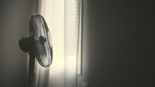 Weißer Standventilator steht in einem abgedunkelten Raum am Fenster