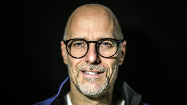 Nahaufnahme des lächelnden Patrick Schubert mit schütterem Haarwuchs, Brille und Bart vor dunklem Hintergrund.