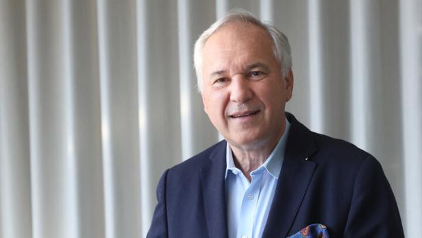 Walter Rosenkranz lehnt an einem Tisch vor hellen Vorhängen und lächelt.