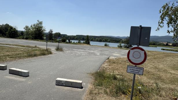 Sperrschilder blockieren eine Straße am Seeufer mit Blick auf Wasser und Hügel.