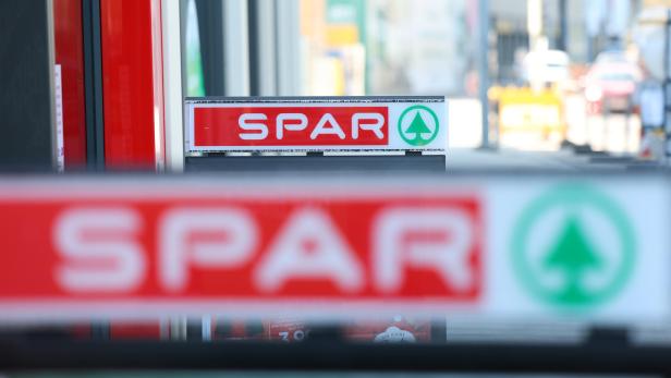 Das Logo der Supermarktkette SPAR ist auf einem Schild vor einem Geschäft zu sehen, wobei ein weiteres Logo im Vordergrund unscharf dargestellt ist.