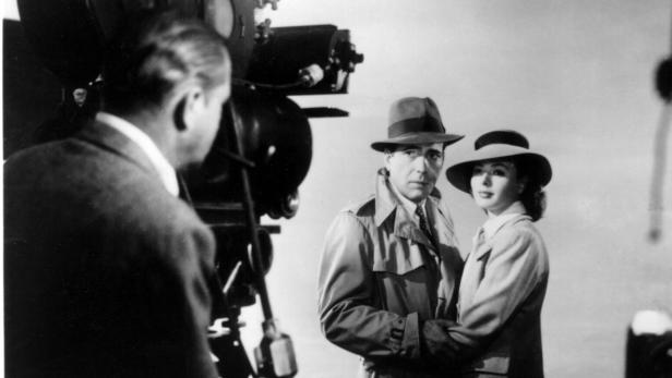 Humphrey Bogart und Ingrid Bergman stehen in Trenchcoats und Hüten vor einer Filmkamera.