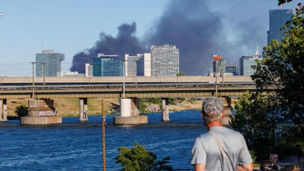 Eine Person blickt von einem Flussufer auf eine dunkle Rauchwolke über der Skyline der Wiener Donau City.