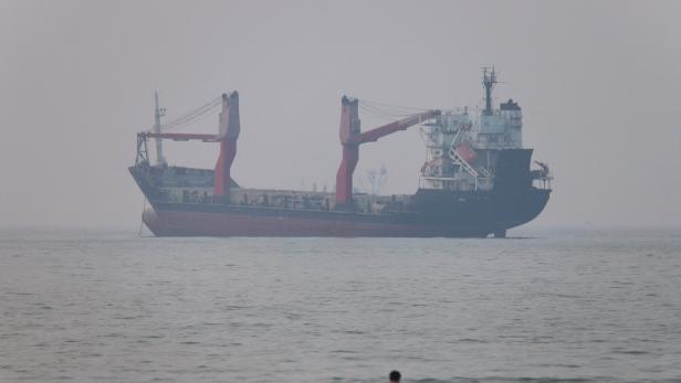 Ein Frachtschiff liegt vor der Küste im Wasser, während eine Gruppe von Menschen im Vordergrund im Meer steht.