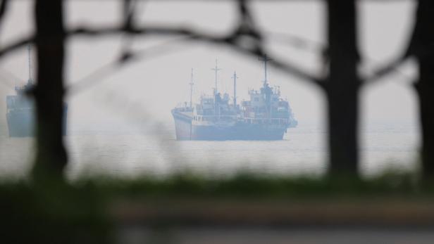 Zwei Frachtschiffe liegen im Nebel auf ruhigem Meer, durch dunkle Gitter im Vordergrund gesehen.
