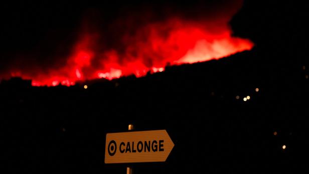 Ein Wegweiser nach Calonge vor einem nächtlichen Waldbrand, der den Himmel leuchtend rot färbt.