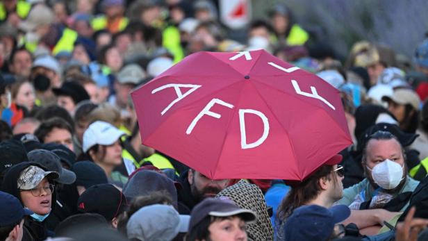 Dichtes Gedränge mit vielen Menschen, im Vordergrund ein roter Regenschirm mit weißen Buchstaben.