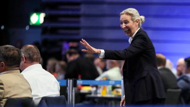 Alice Weidel lacht und winkt in einer Menschenmenge vor blauem Hintergrund.