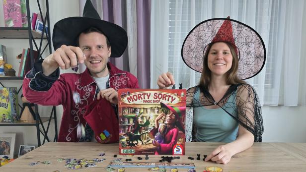 Ein Paar in Hexenkostümen spielt das Brettspiel „Morty Sorty Magic Shop“ an einem Holztisch.