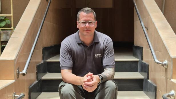 Christoph Riedl mit Brille und Tattoo sitzt auf einer Treppe vor einem dunklen Eingang.