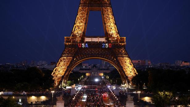 Der beleuchtete Eiffelturm in Paris zeigt bei Nacht die Aufschrift „USA 250“ über einer belebten Straße.