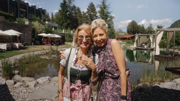 Zwei Frauen lächeln vor einem Naturbadesee mit Bühnenzelt im Alpenresort.