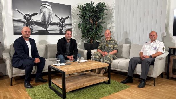 Vier Männer sitzen in einem hellen Sitzbereich auf Sofas um einen Couchtisch, dahinter hängt ein Flugzeugbild.