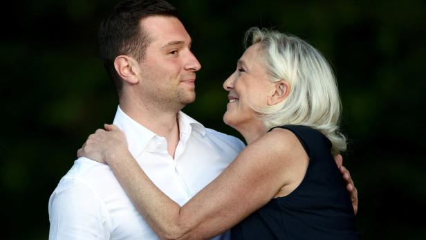 Marine Le Pen lächelt Jordan Bardella an, während sie ihn vor einem dunklen Hintergrund umarmt.