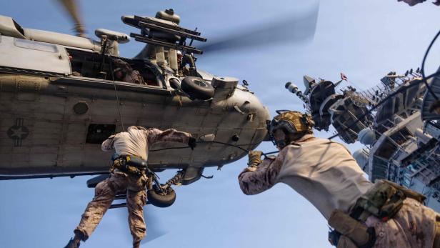 Ein Soldat seilt sich von einem Hubschrauber auf das Deck eines Kriegsschiffes ab, während ein Kamerad die Aktion sichert.