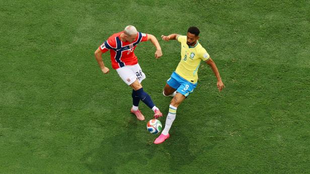 Ein norwegischer Fußballspieler im roten Trikot kämpft auf dem Rasen mit einem brasilianischen Spieler im gelben Trikot um den Ball.