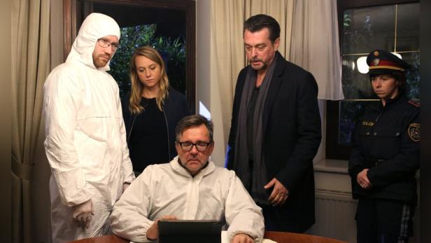 Ein Ermittlerteam und eine Polizistin beobachten einen Mann in Schutzkleidung, der an einem Tablet arbeitet.