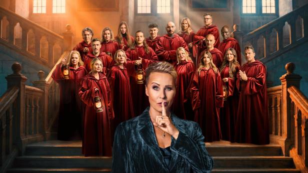Sonja Zietlow steht im Vordergrund und macht eine Schweigegeste vor einer Gruppe in roten Umhängen auf einer Treppe in der Show „Die Verräter“.
