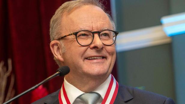 Der australische Premierminister Anthony Albanese lächelt vor einem Mikrofon und trägt ein rot-weißes Ordensband um den Hals.
