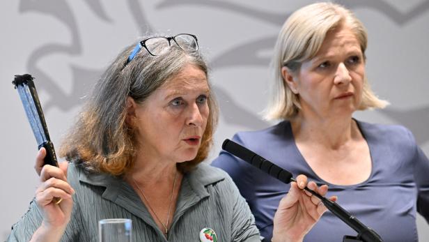 Zwei Frauen bei einer Pressekonferenz; die vordere spricht ins Mikrofon und hält einen Gegenstand hoch.