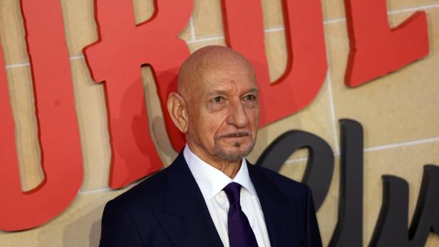 Ben Kingsley trägt einen dunklen Anzug mit weißem Hemd und violetter Krawatte vor einem Hintergrund mit großen roten Buchstaben.