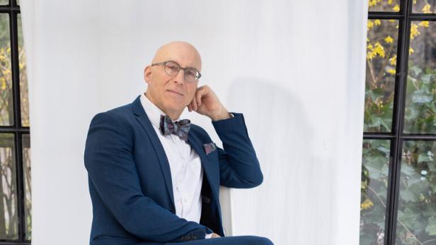 Ein glatzköpfiger Mann mit Brille und Fliege sitzt in einem blauen Anzug auf einem weißen Stuhl vor einem hellen Hintergrund.