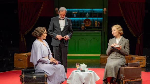 Auf einer Theaterbühne sitzen zwei Frauen mit Koffern an einem Tisch, während ein Mann im Anzug zwischen ihnen steht.
