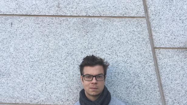 Ein junger Mann mit Brille und Schal steht vor einer hellen, gesprenkelten Steinwand.