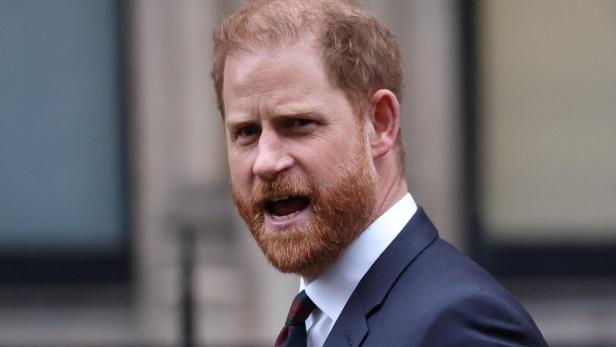 Prinz Harry trägt einen dunklen Anzug mit Krawatte und spricht mit offenem Mund vor einem unscharfen Hintergrund.