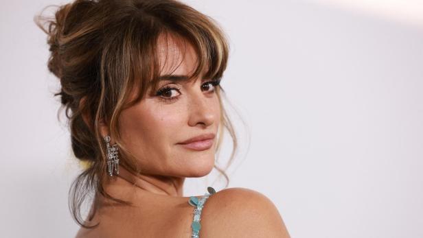 Penélope Cruz blickt mit hochgestecktem Haar und eleganten Ohrringen über die Schulter in die Kamera.