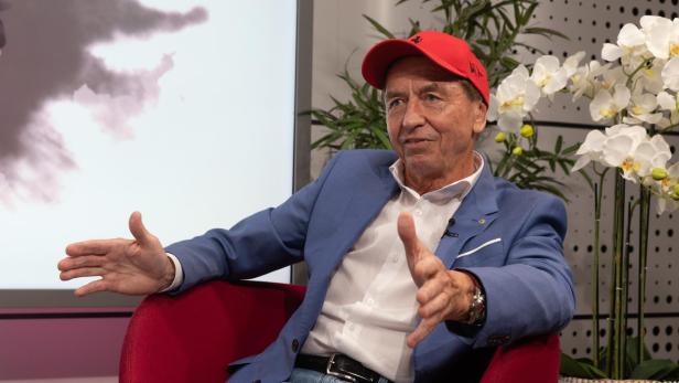 Heribert Kasper sitzt in einem roten Sessel, trägt ein blaues Sakko sowie eine rote Kappe und gestikuliert während eines Gesprächs.