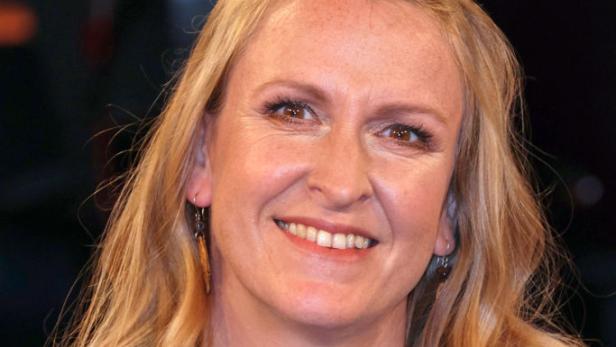 Eine lächelnde Frau mit blondem Haar und Jeansjacke vor dunklem Hintergrund.