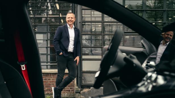 Mann im blauen Blazer steht lächelnd vor einem Auto und einem Industriegebäude.