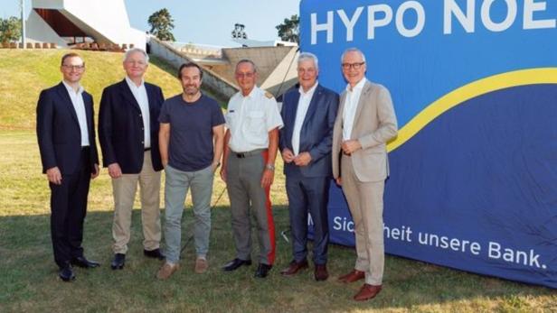 Sechs Männer stehen vor einer großen blauen HYPO NOE-Wand auf einer Wiese.