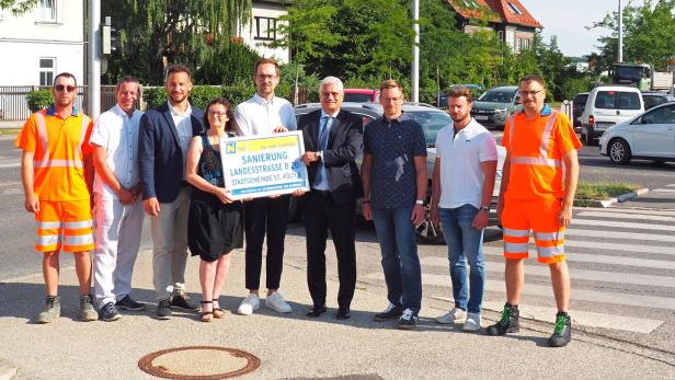 Gruppe von neun Personen an einer Kreuzung hält ein Schild zur Sanierung der Landesstraße B20 in St. Pölten.