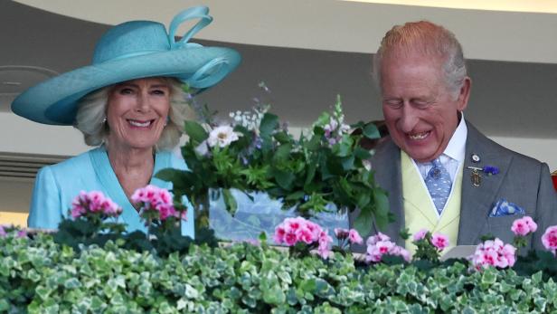 König Charles III. und Königin Camilla lächeln gemeinsam hinter einem mit Blumen geschmückten Balkon.