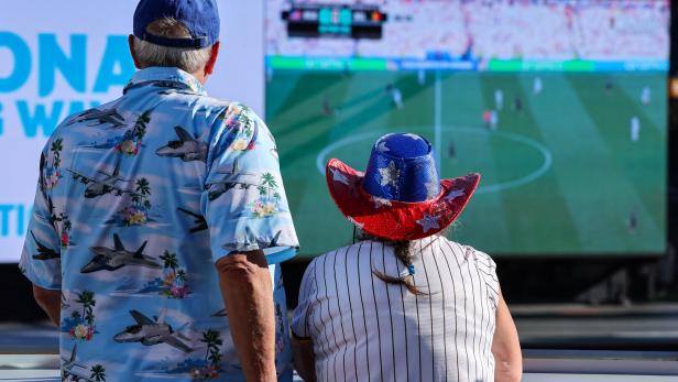 Zwei Personen mit patriotischen Hüten schauen ein Fußballspiel auf einer großen Leinwand.
