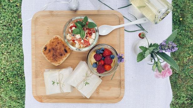 Ein Picknick auf einer Wiese mit einem Holzbrett, das belegte Sandwiches, einen Tomaten-Feta-Salat, ein Beeren-Dessert und Sekt umfasst.