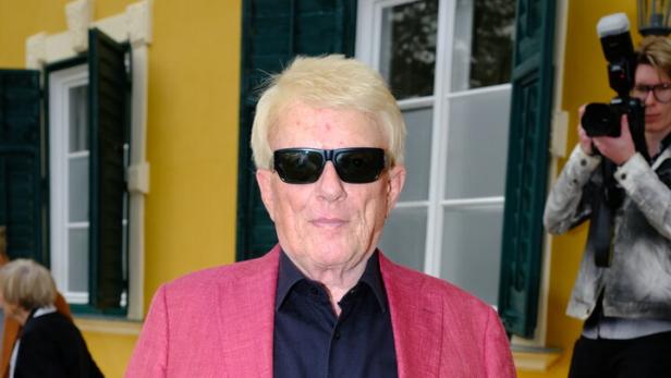 Heino trägt ein pinkes Sakko, ein schwarzes Hemd und eine Sonnenbrille vor einem gelben Gebäude.