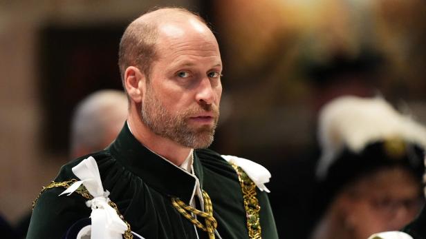 Prinz William trägt in zeremonieller Kleidung mit grünem Umhang und goldener Kette ein Programmheft in den Händen.