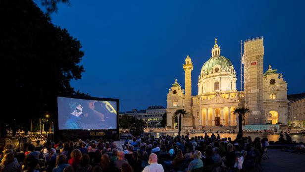 Menschen sitzen bei einer Freiluftfilmvorführung vor einer beleuchteten Barockkirche am Abend.