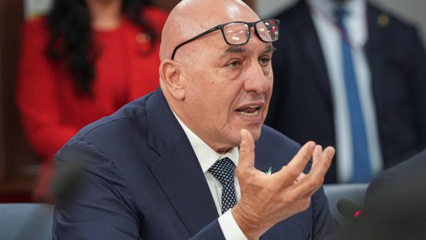 Ein Mann im Anzug spricht bei einer Sitzung und gestikuliert mit der Hand, während eine Brille auf seinem Kopf sitzt.