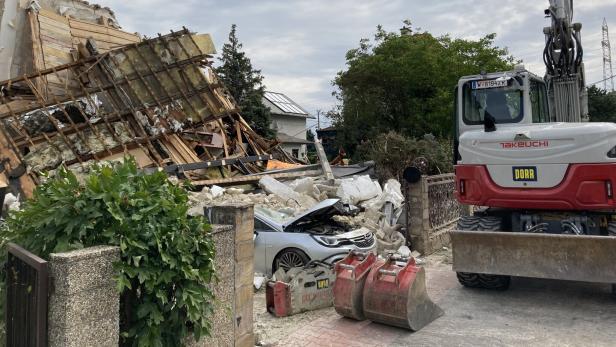 Abrissstelle mit eingestürztem Haus, verschüttetem Auto und Bagger neben Trümmern.