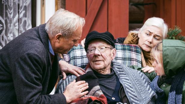 Älterer Mann sitzt eingehüllt im Sessel, umgeben von besorgten Angehörigen draußen vor einem Haus.